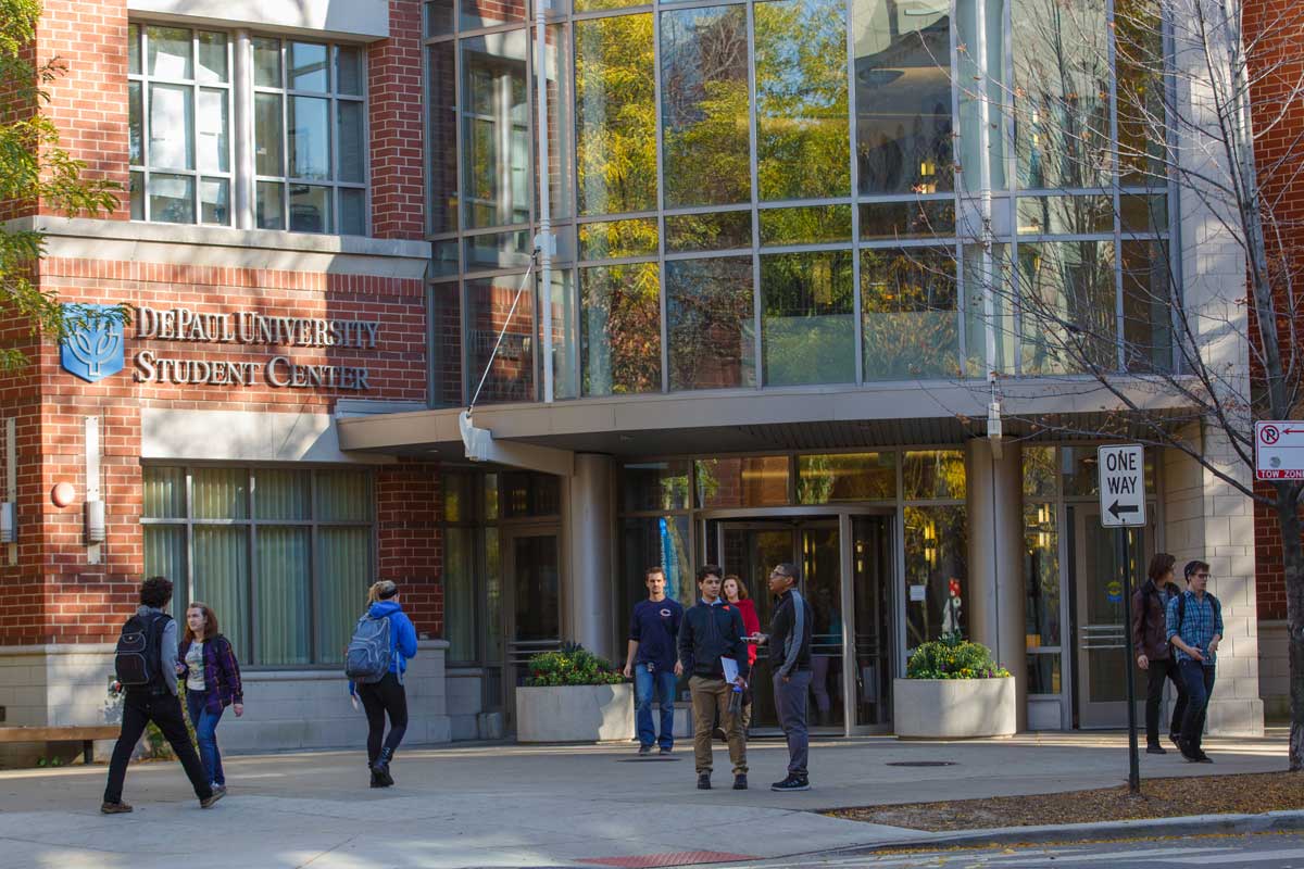 Students stand outside the entrance of a tall modern building with glass front. 