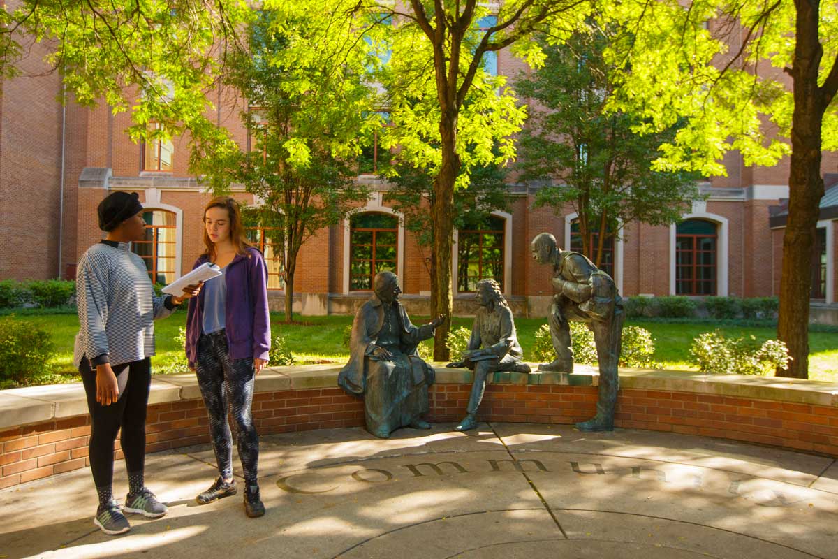 Two female students standing in a courtyard, looking at a notebook. 
